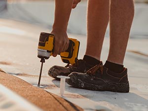 Flat-Roof-Repair-McKinney-TX-Texas-1 | Anderson Industrial Roofing Person in sneakers drives a screw into a wooden surface with a yellow cordless drill outdoors, close to the ground.