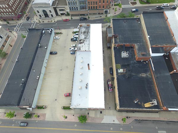 Aerial view of a downtown block with flat-roofed buildings, a white central roof, parked cars, and surrounding streets.