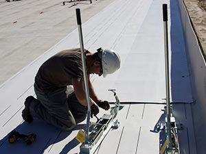 Roof-Maintenance-McKinney-TX-Texas-21 | Anderson Industrial Roofing Construction worker in a white hard hat kneeling on a white roof, fastening metal brackets with a drill between two vertical posts.