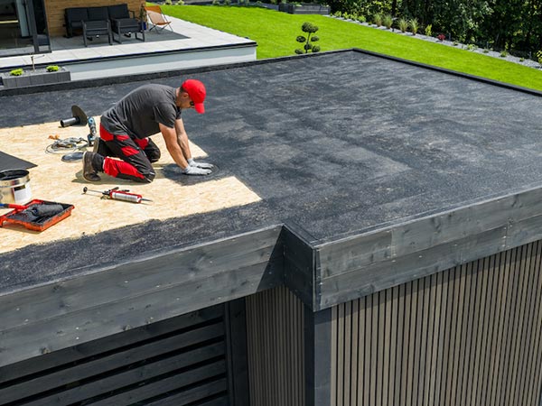 Roof-Maintenance-Grand-Prairie-TX-Texas-2 | Anderson Industrial Roofing Man wearing a red cap kneels on a flat roof, patching a black roofing membrane with tools nearby.