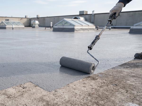 Roof-Restoration-Grand-Prairie-TX-Texas-2 | Anderson Industrial Roofing Roofer applying gray waterproof coating to a flat roof with a paint roller, wearing gloves.