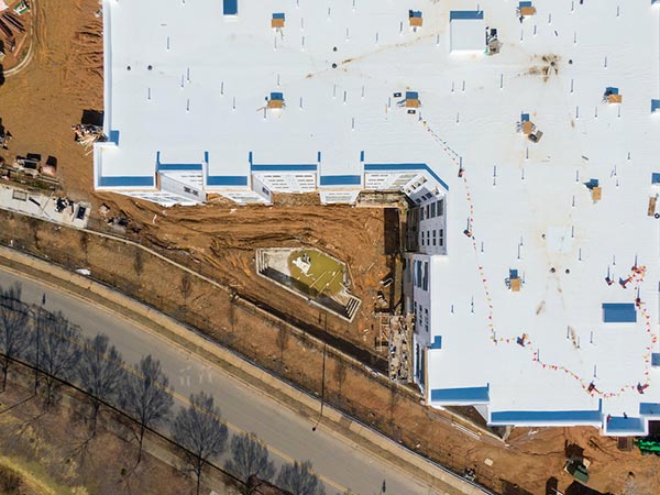 Aerial view of a large white-roofed building under construction, with exposed dirt around the base and a rooftop excavation near the corner beside a road.
