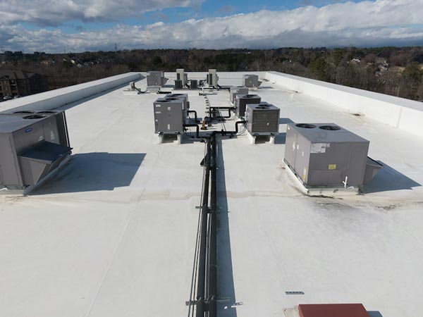 Wide rooftop with multiple rooftop HVAC units and supporting pipes, extending toward a tree line under a cloudy sky.